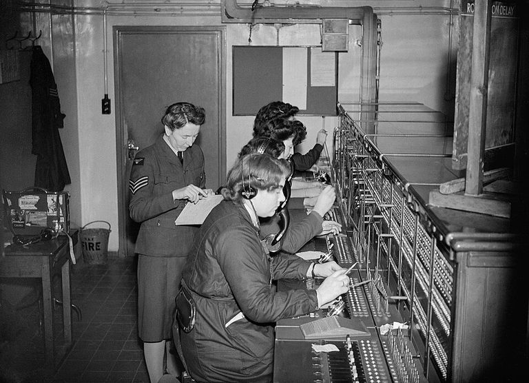 Three WAAF telephone operators in headsets patching calls on a long switchboard while a sergeant takes notes, Uxbridge, 1942