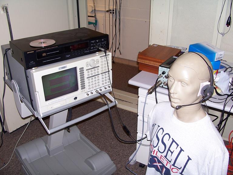 A test mannequin head wearing a call-centre operator headset with a boom microphone, wired to a signal analyzer and CD player in an acoustics lab
