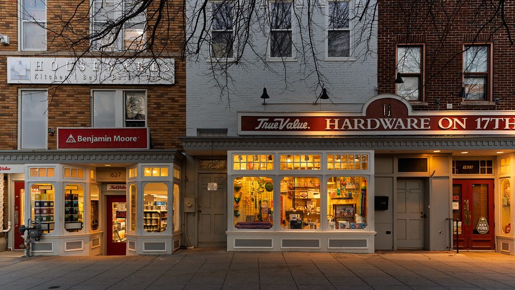Small town storefronts at dusk, lights on behind the windows of a hardware shop