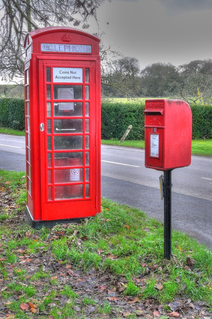 A red British telephone box on a village verge with a sign reading Coins Not Accepted Here, next to a red post box
