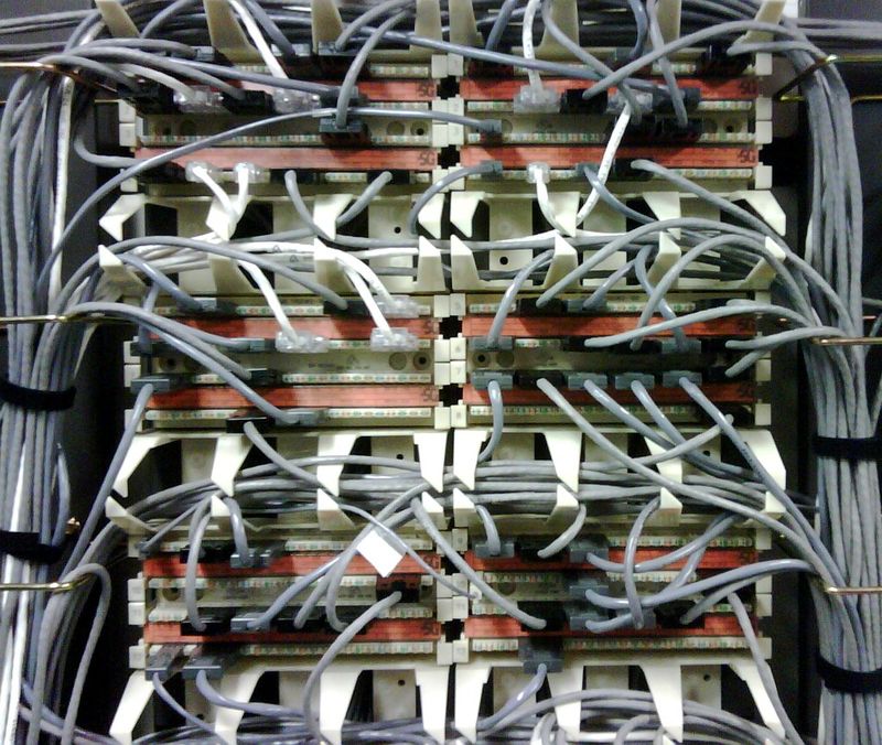 Telecom patch panel with dozens of grey network cables plugged into rows of punch-down blocks and looping off to bundles on both sides