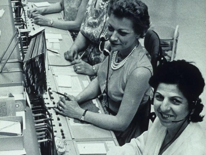 Telephone switchboard operators wearing headsets sit at a long console of cords, keys and jacks, routing calls