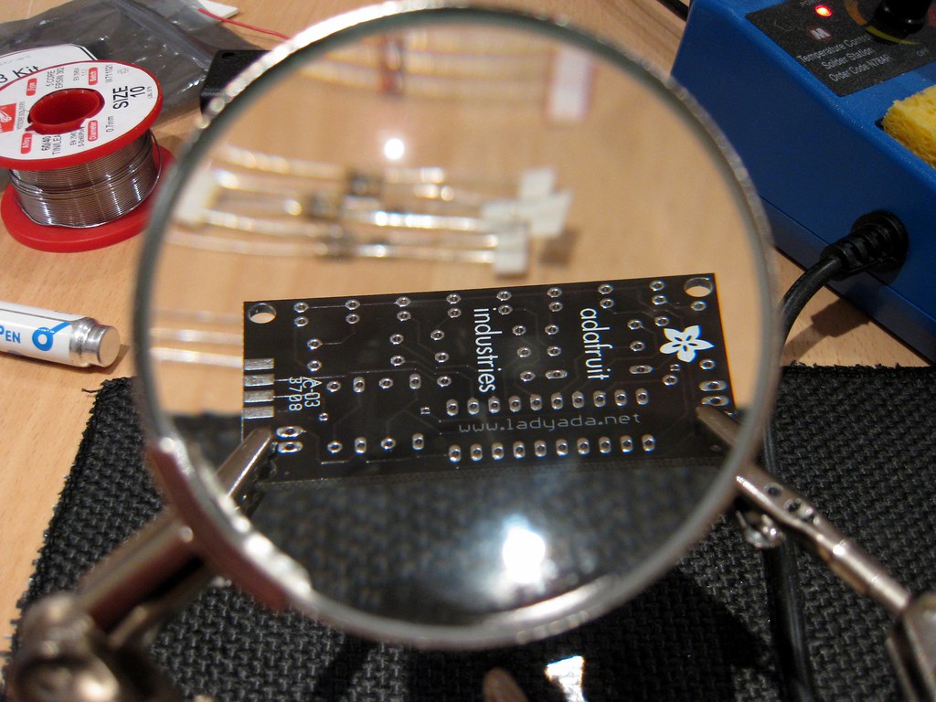 Magnifying glass held over a bare circuit board on a workbench, next to a spool of solder and a soldering station