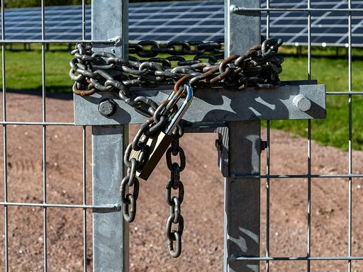 A heavy chain and brass padlock wrapped around the gate of a fenced solar park, with rows of solar panels behind the wire mesh