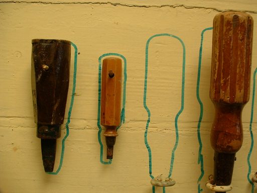 Old hand tools hung on a yellow workshop wall, each over its own painted outline, with one outline left empty where a tool is missing