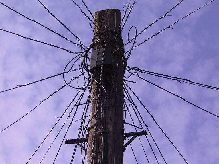 A wooden telephone pole seen from below, with dozens of wires radiating out from the top against a cloudy sky